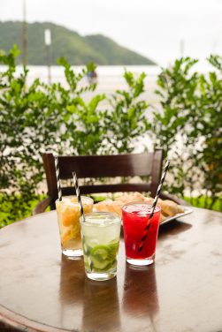 Três diferentes tipos de caipirinha de frutas sobre mesa externa da Churrascaria Tertúlia com vista para o mar