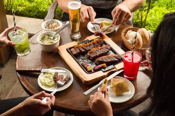 Mesa externa da Churrascaria Tertúlia com uma porção de tiras de carne na chapa, petiscos, acompanhamentos e drinks.