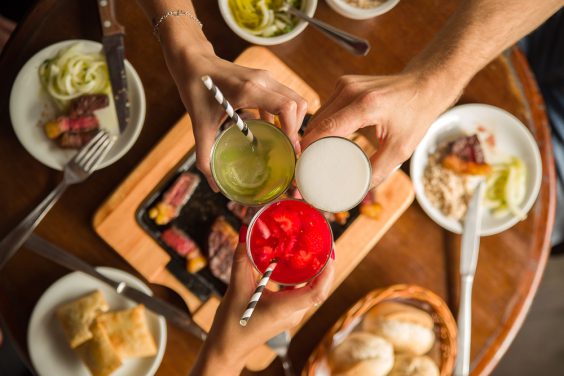 Brinde com chopps e caipirinhas sobre mesa servida com chapa de carnes, pastéis, pães e outros acompanhamentos